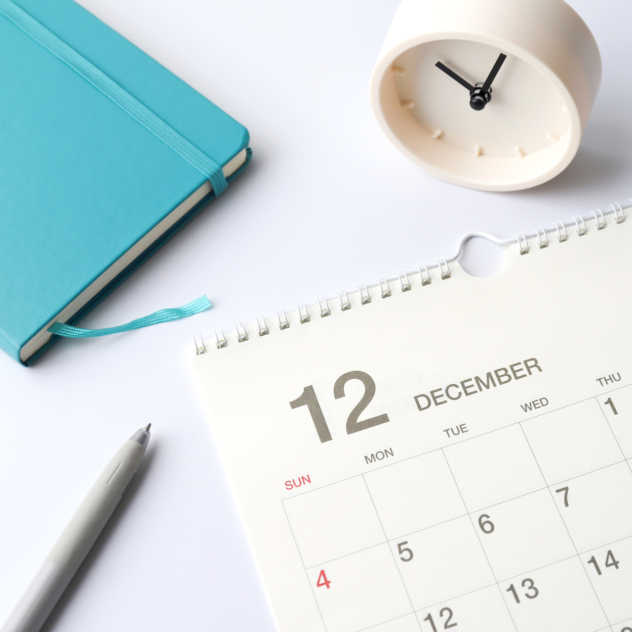 12月のカレンダー、青い手帳、白いアナログ時計、ペンが並んだデスクの上の平置き構成。予定管理やスケジュール確認をイメージさせる写真。